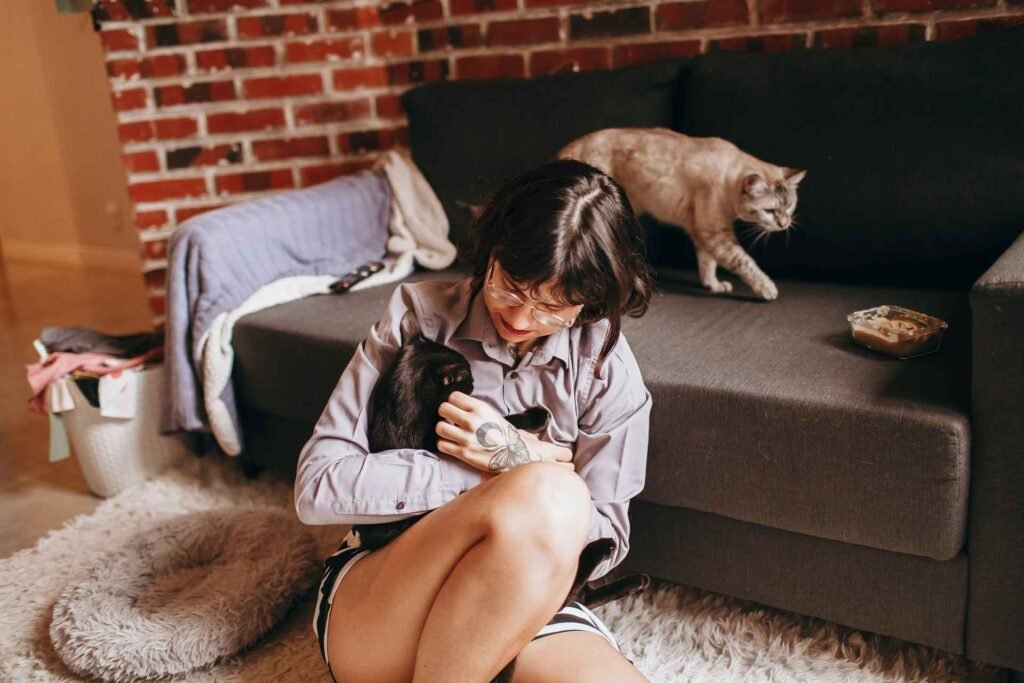 A woman with a black cat in her arms and an orange cat behind her walking on a couch.